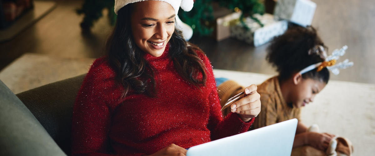 Woman in holiday sweater shopping on her laptop with daughter in background