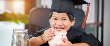 Young boy wearing a graduation cap holding a piggy bank and a jar full of coins.