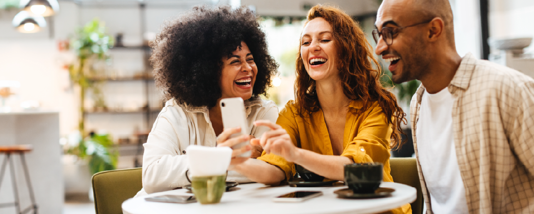 Two women and one man sitting around a table at a coffee shop, laughing and looking at a cell phone together.